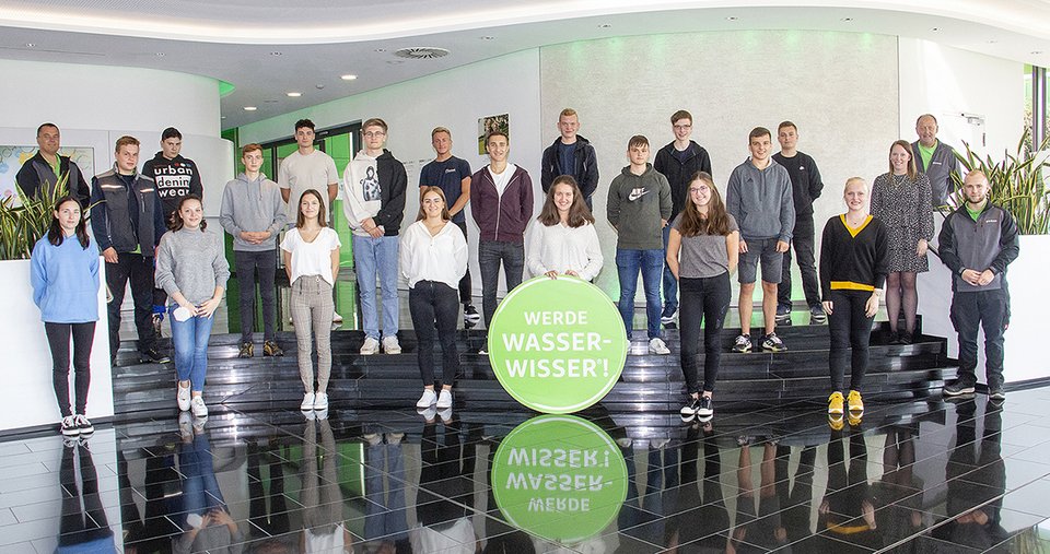 Gruppe von Personen steht auf schwarzem Podest vor weißen Wänden; im Vordergrund ein grünes Schild mit der Aufschrift "WERDE WASSERWISSER!"