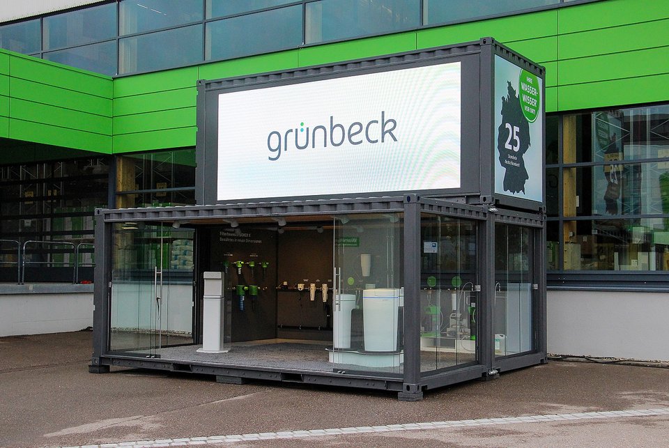 Ein modularer Container mit der Aufschrift "grünbeck" steht vor einem Gebäude, innen befinden sich verschiedene technische Ausstellungsstücke.