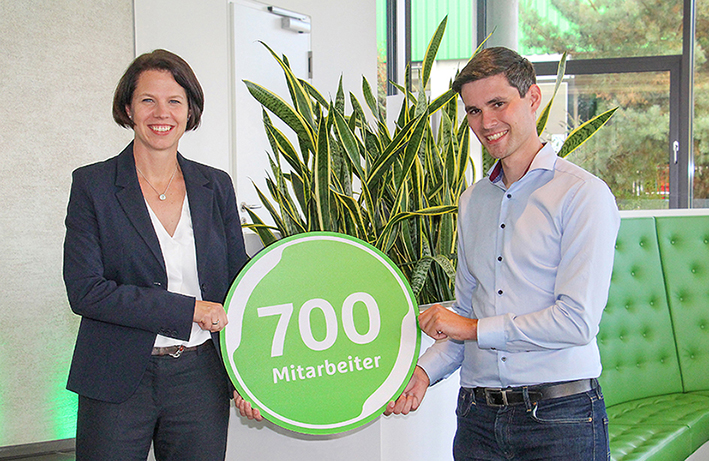Zwei Personen halten ein rundes, grünes Schild mit der Aufschrift "700 Mitarbeiter" in einem Büro vor einer Pflanze.