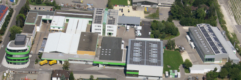 Großer Industriekomplex mit mehreren Gebäuden und Photovoltaikanlagen auf den Dächern, umgeben von grüner Vegetation und Parkplätzen.
